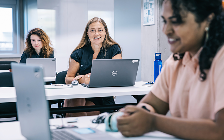 On the right is a woman sitting in a classroom looking at a laptop. Her image is slightly blurred. Behind her are two women smiling and looking at laptops.