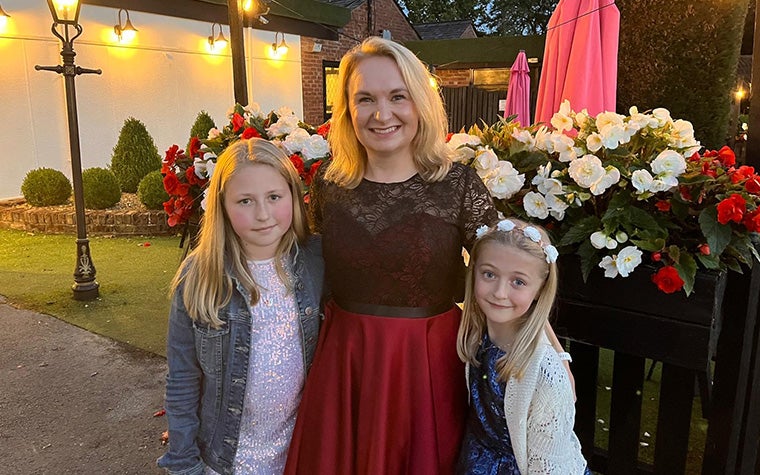 A woman (Joanne Staples) is smiling at the camera and standing in between her two daughters.