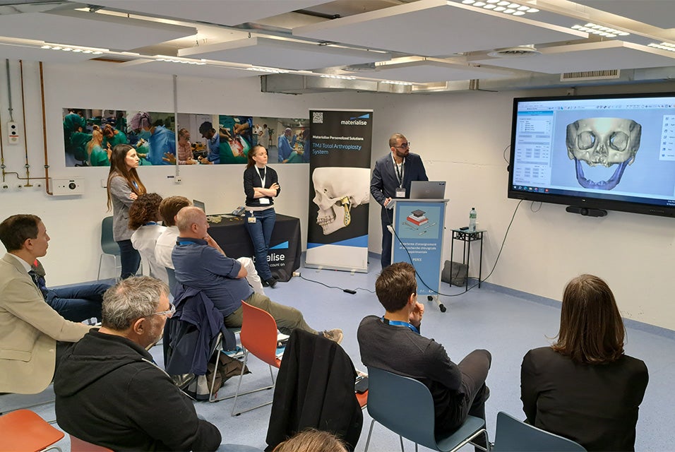 A presenter speaking to a seated audience during a Materialise CMF Wet Lab session, with a large screen displaying a 3D skull model within Materialise Mimics Enlight CMF software.