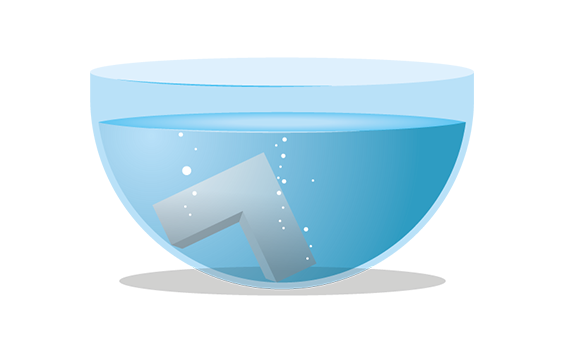 A gray L-shaped object submerged in a transparent bowl filled with blue liquid, with small air bubbles rising around it.