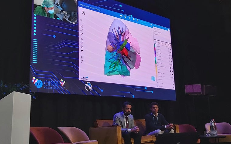 Two men sitting on stage with microphone headsets during the Orsi Surgical AI Days conference. Above them is a large screen displaying the OR where the surgery is taking place and a screenshare of the Materialise Mimics software used to plan the surgery.