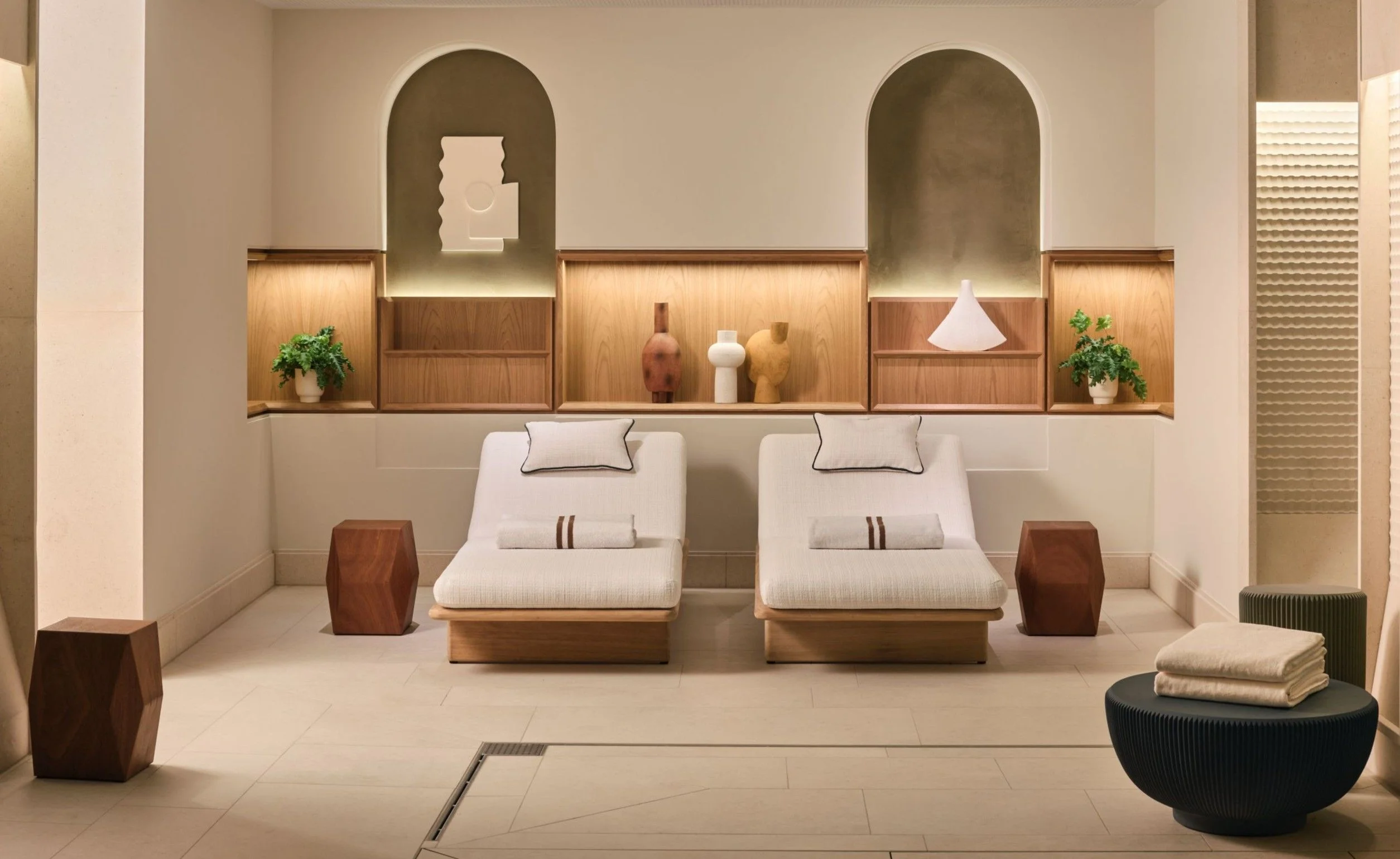Minimal spa relaxation area with two upholstered loungers, wooden details, arched wall niches and warm ambient lighting, interior design by Erlarcher.