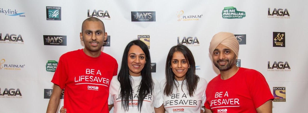 Four DKMS volunteers wearing branded t-shirts in front of a banner 