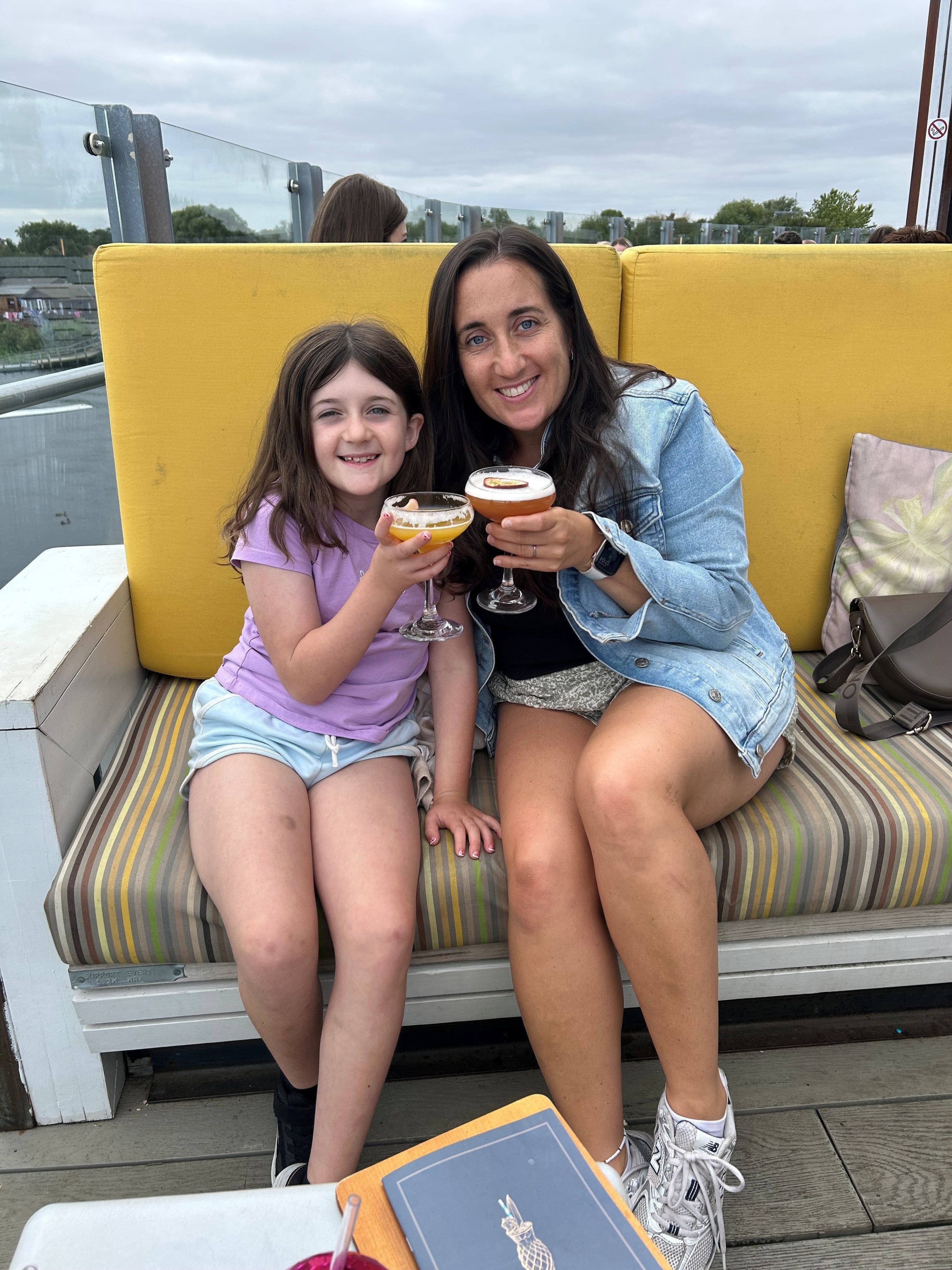 Little Girl and her mum sitting outside on a couch.  They are "cheers"ing their drinks and smiling. 