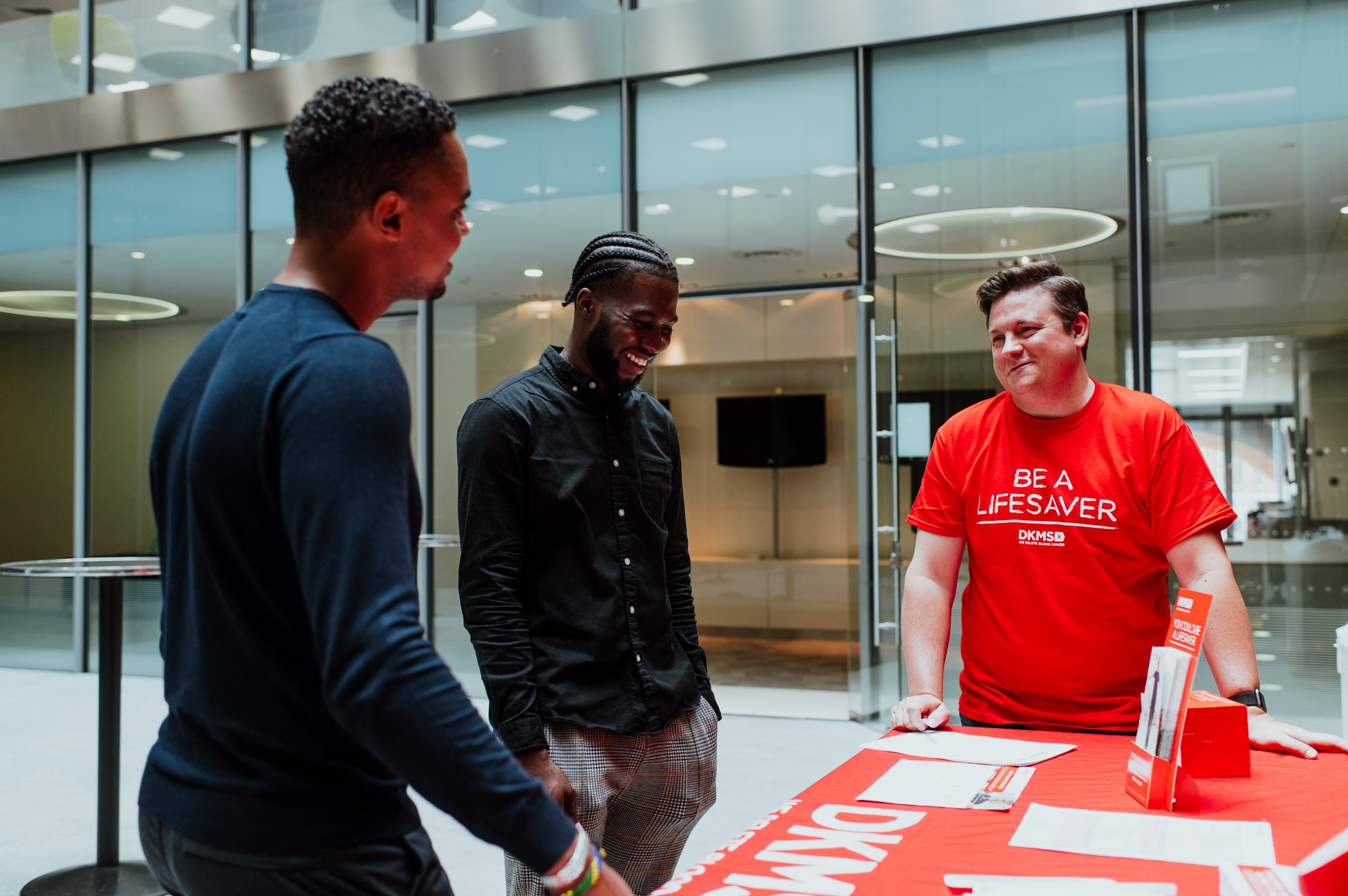 Three men gathered around a table, one wearing a DKMS t-shirt