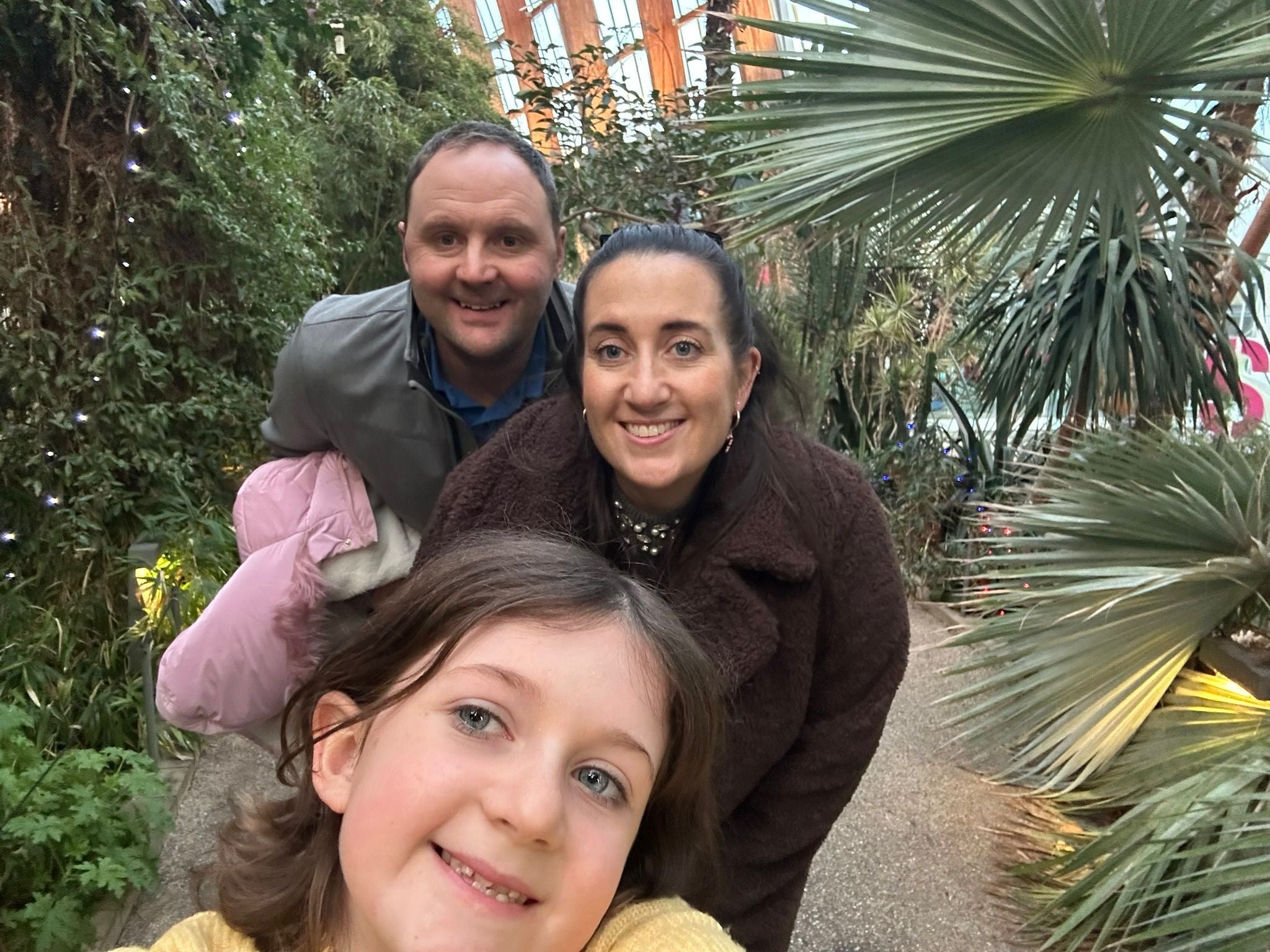 A dad, mum and their daughter smile in a selfie while in a botanical garden.  