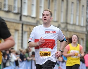 Man wearing DKMS Top runs in a race, with other runners and supporters in the background. 