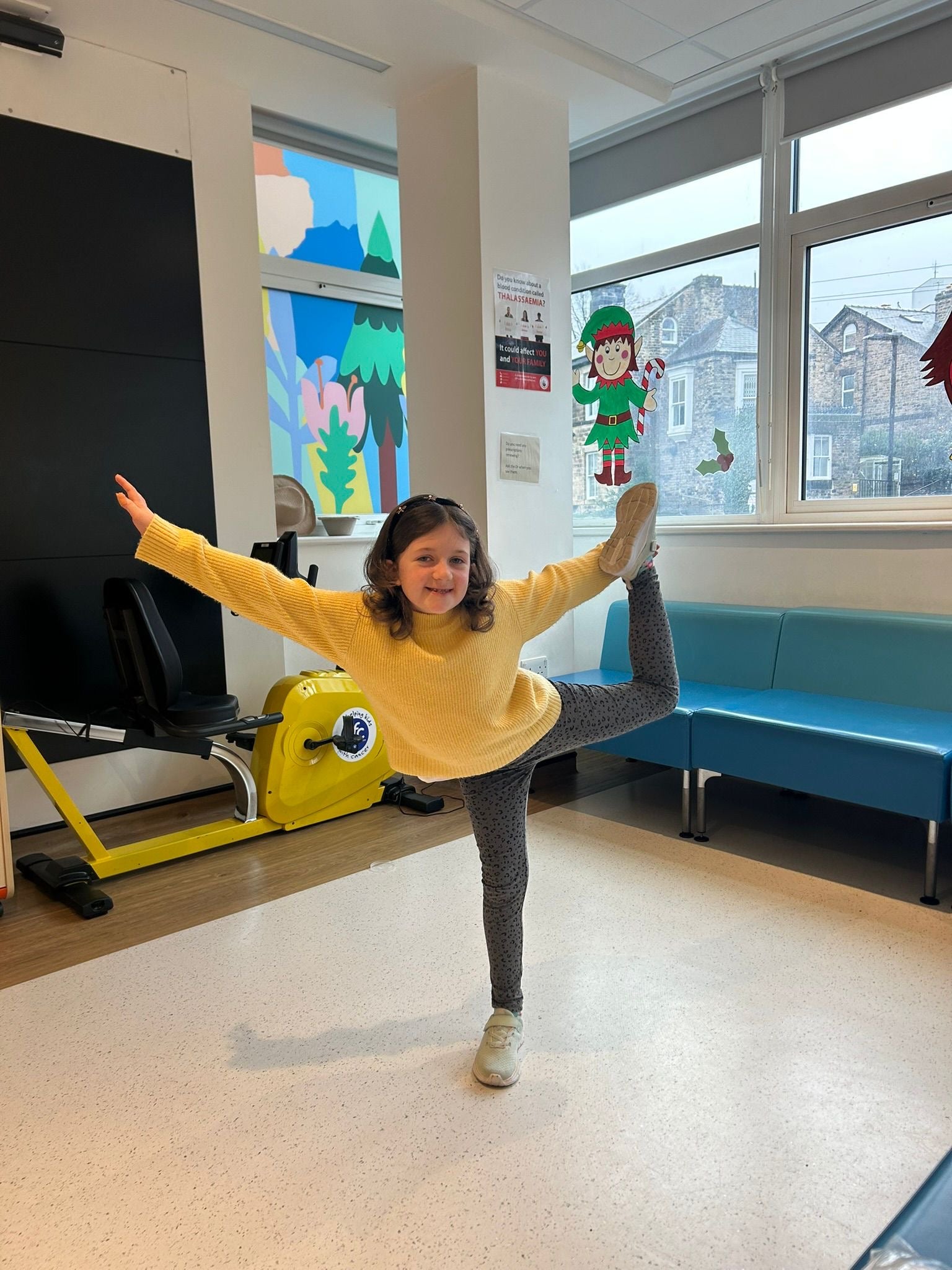A little Girl dances in a waiting area.  She is smiling and standing on one leg. 