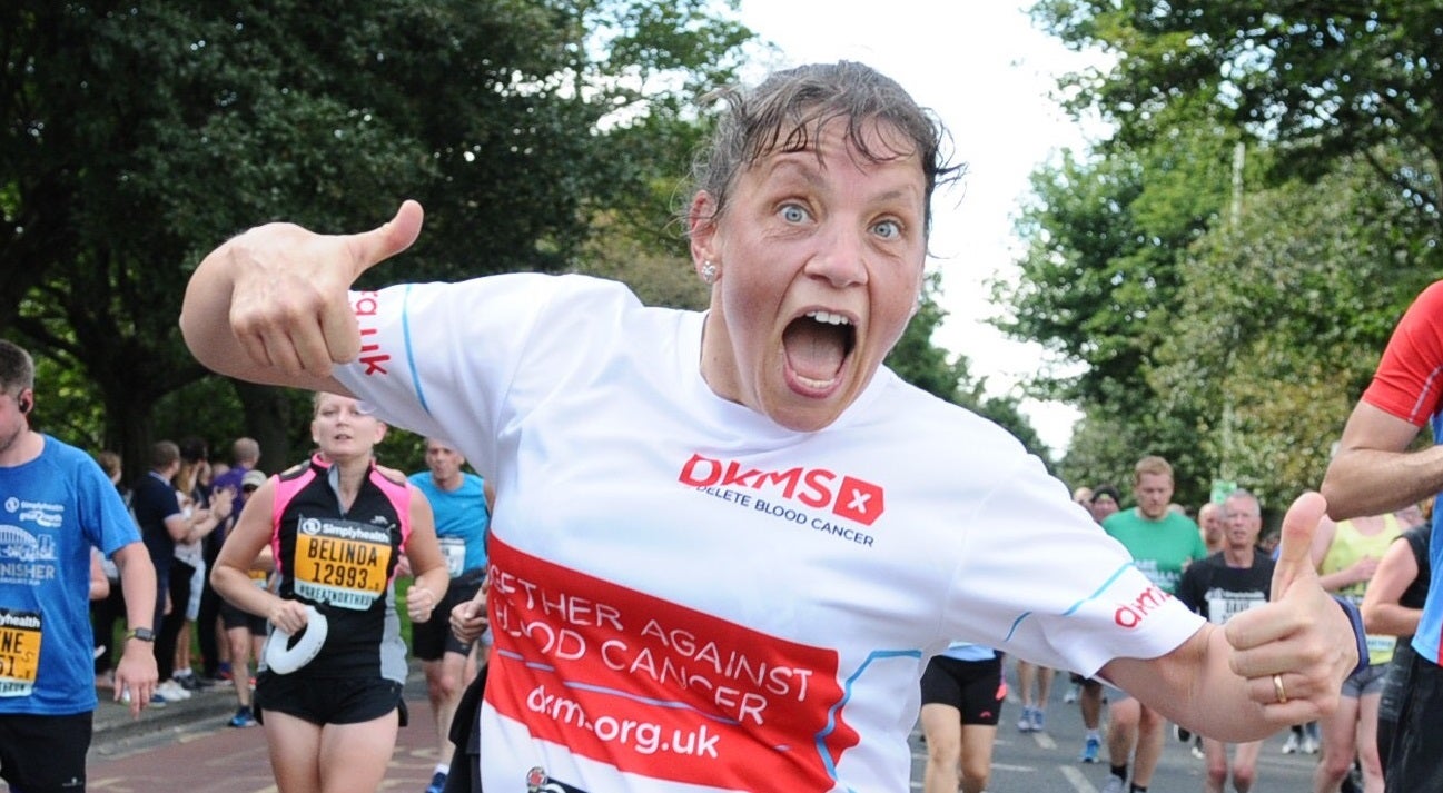 Woman wearing DKMS running top holds two thumbs up while running race, with other runners and supporters in the background.