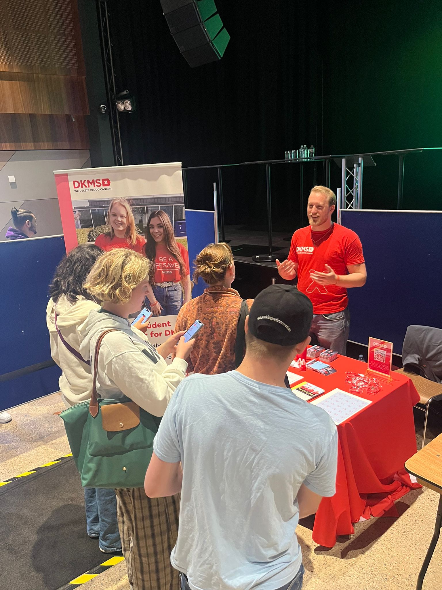 Students speaking to Middle aged White man at a DKMS branded stand