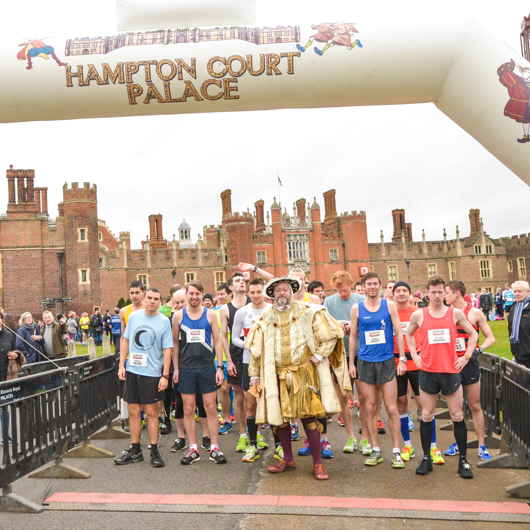 A man dressed as Henry VIII stands at the start line of the Hampton Court Palace Race, surrounded by runners. 