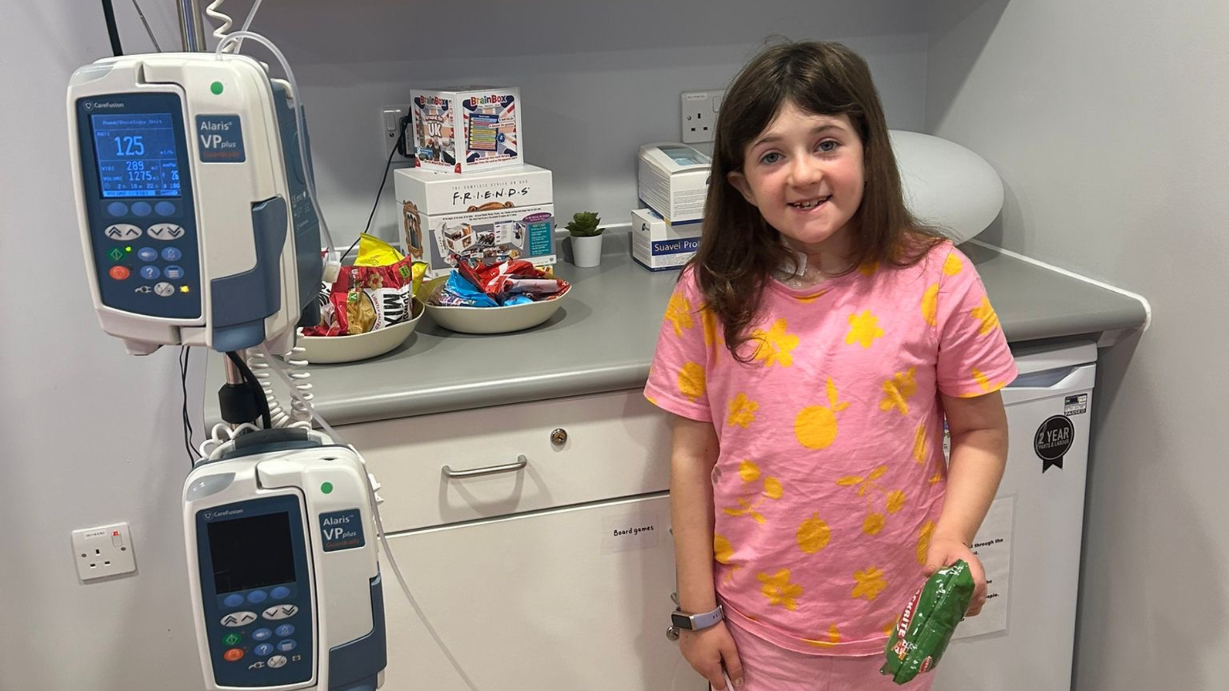 A little girl in hospital stands next to monitors, smiling and holding a bag of crisps. 