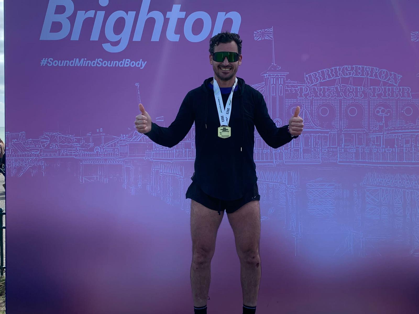 Man stands in front of of Brighton Marathon Banner, with two thumbs up, smiling and wearing a medal. 