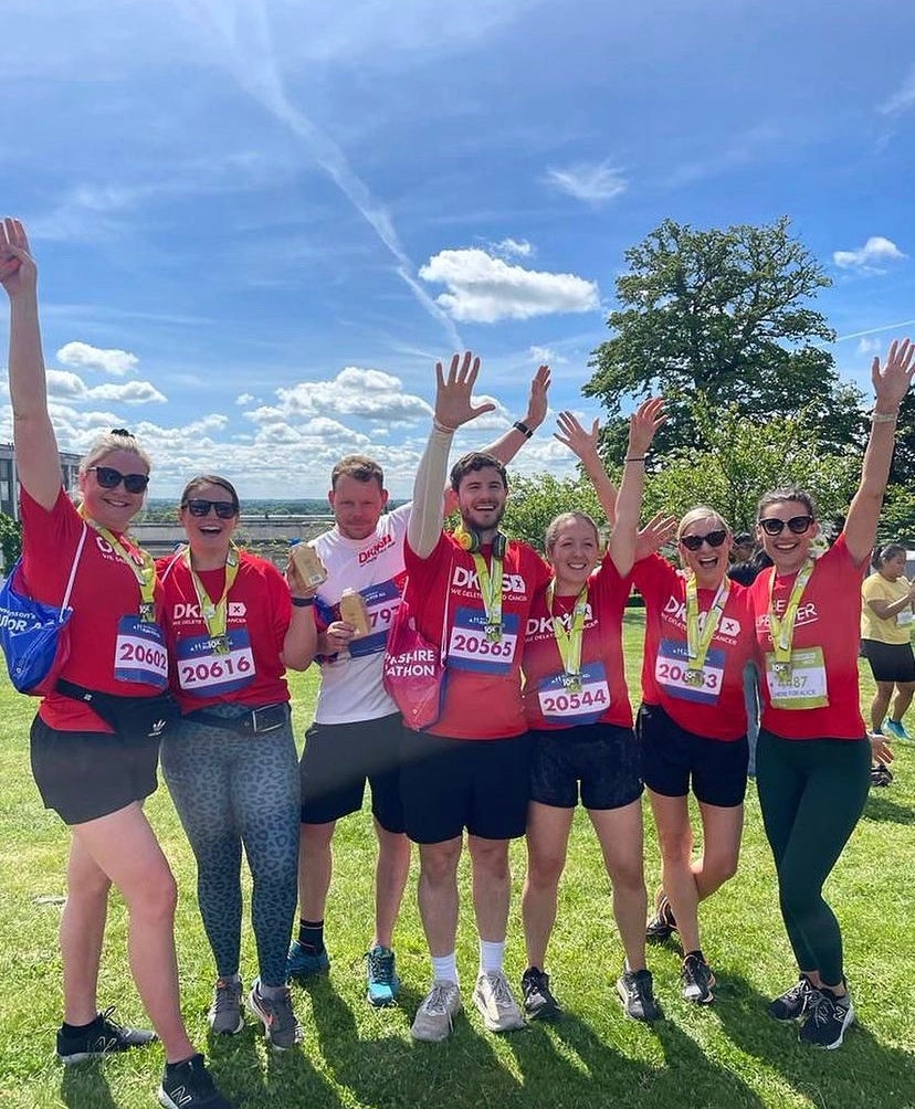 Group of runners stand on grass, holding their hands up in celebration, while wearing Red DKMS tops and race medals. 
