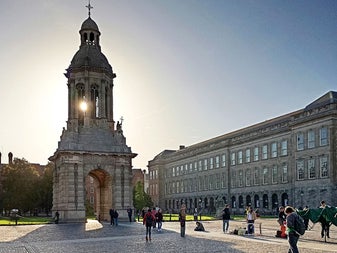 The Campanile at Trinity College Dublin with sunlight behind it and people walking across the cobblestone courtyard