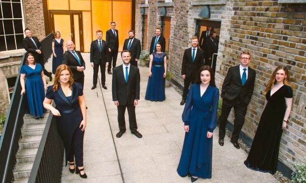 A group of men in dark suits and women in navy, long dresses are standing, scattered around a paved courtyard area.