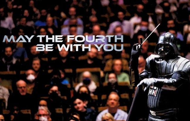 A figure in black shiny darth vadar outfit with a conductor's baton against background of an audience