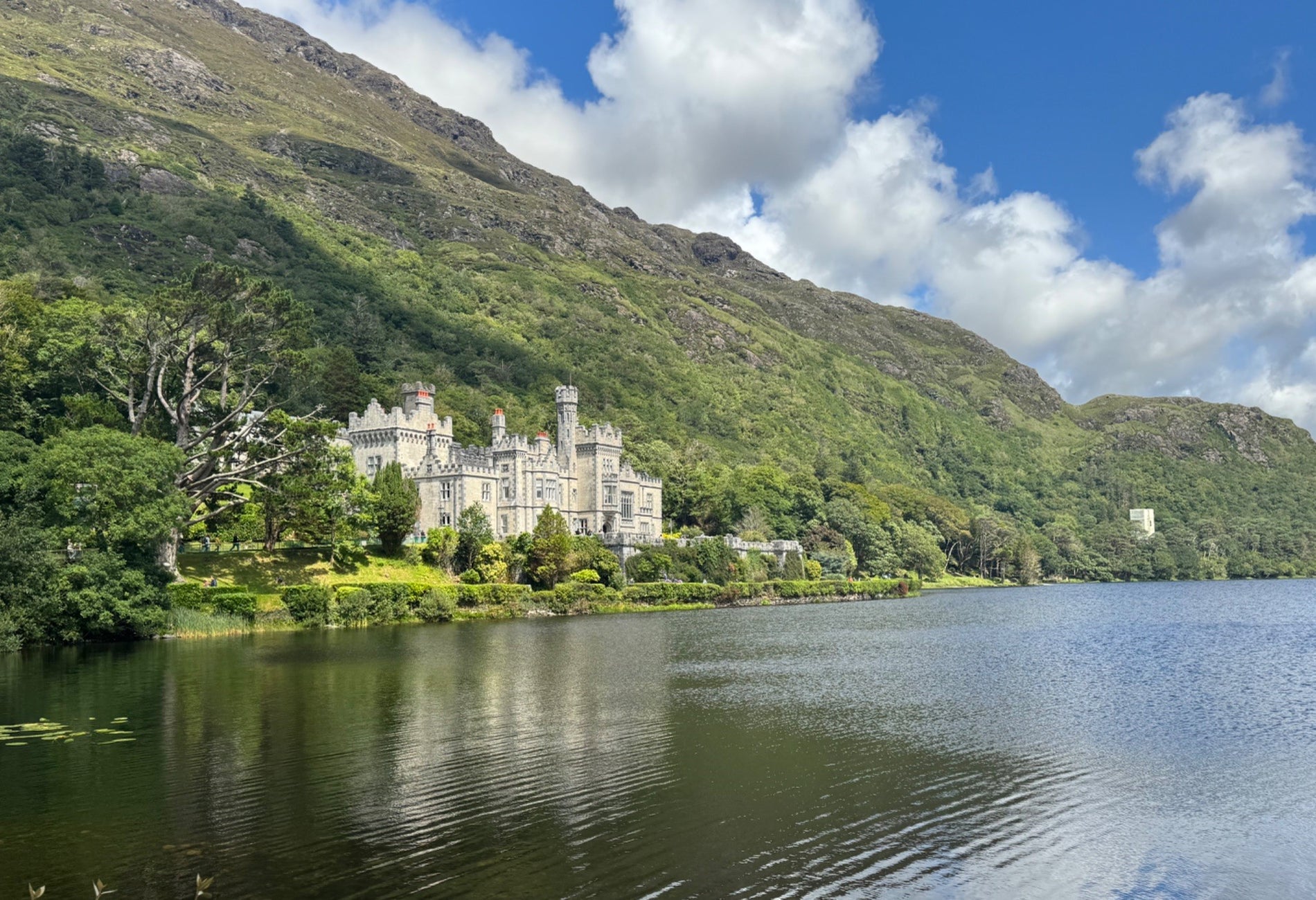 Abbey nestled among trees and mountains with a lake in front of it