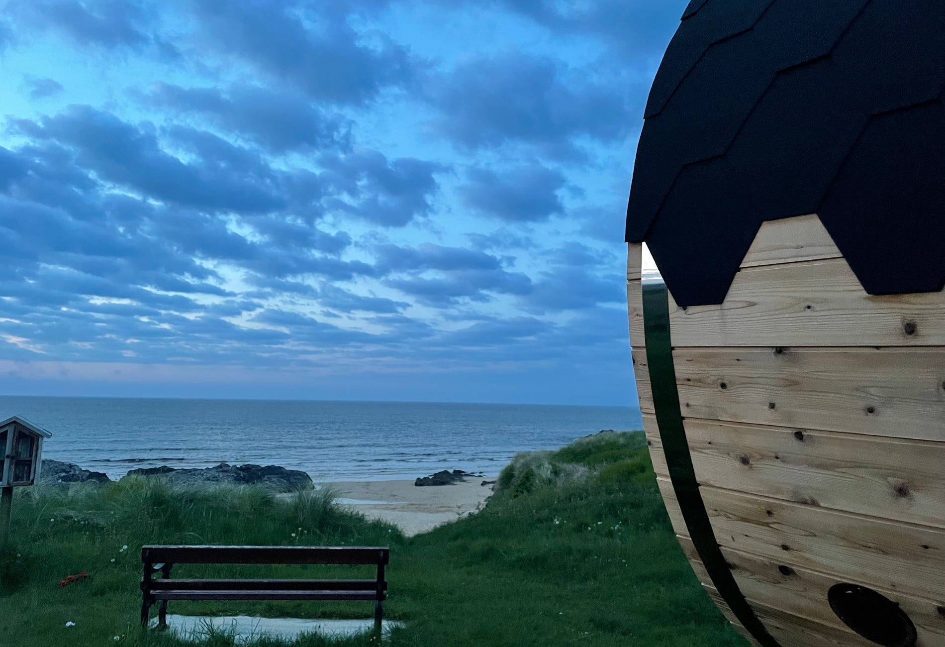 Wooden sauna next to a bench overlooking a beach and grass in the evening time
