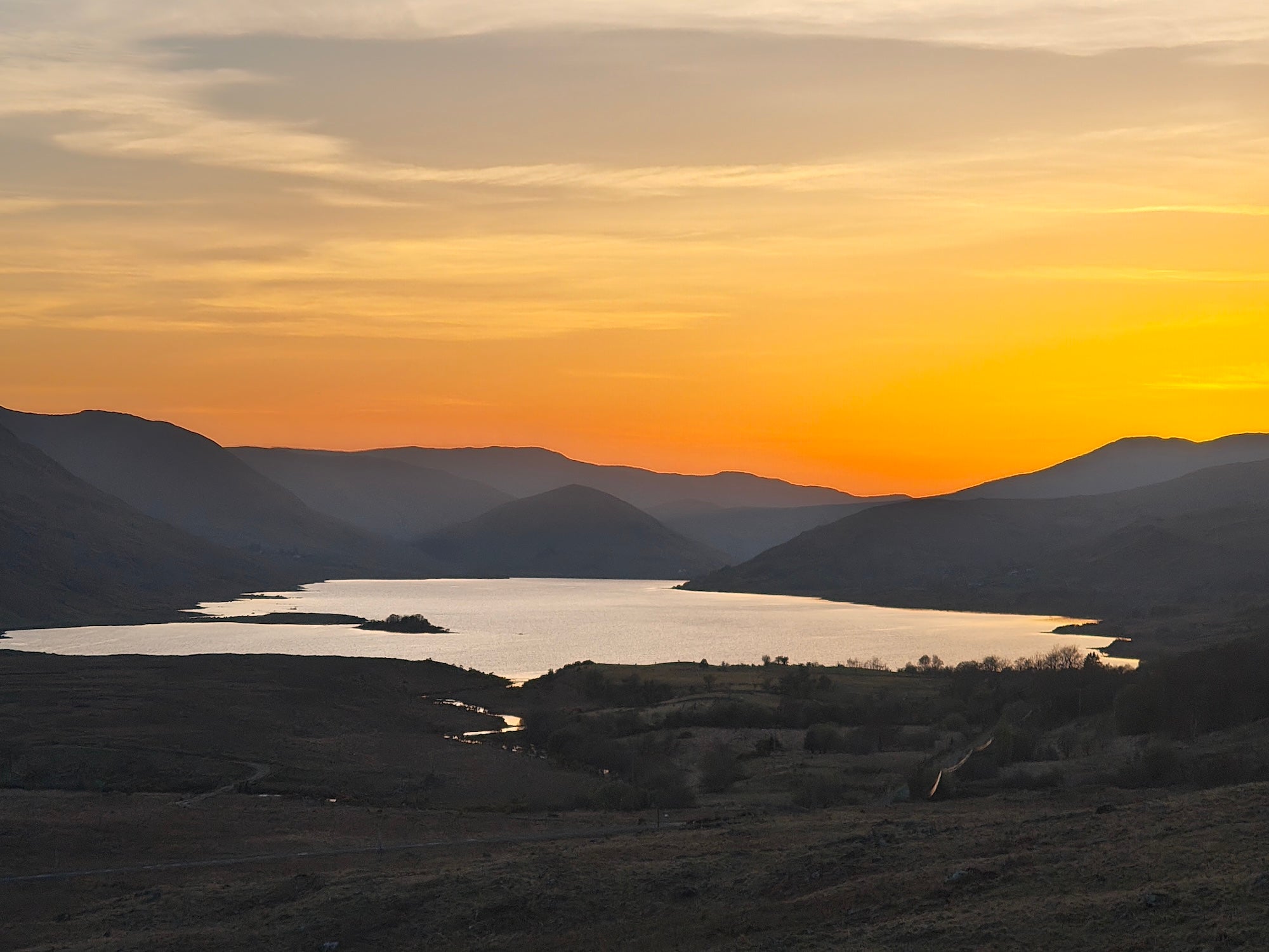 Sunset over Lough Nafooey with golden light reflecting on the lake, surrounded by layered mountain silhouettes and rolling hills in Joyce Country.