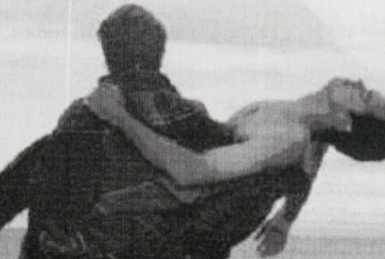 Blurred black and white image of the rear view of a person carrying another person with their head hanging down.