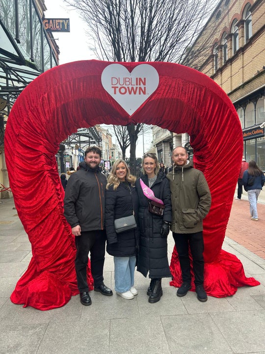Friends pose beneath a giant red velvet heart arch in the heart of Dublin, part of the city's playful embrace of romance and love around Valentine's season, with the Gaiety Theatre visible in the background.