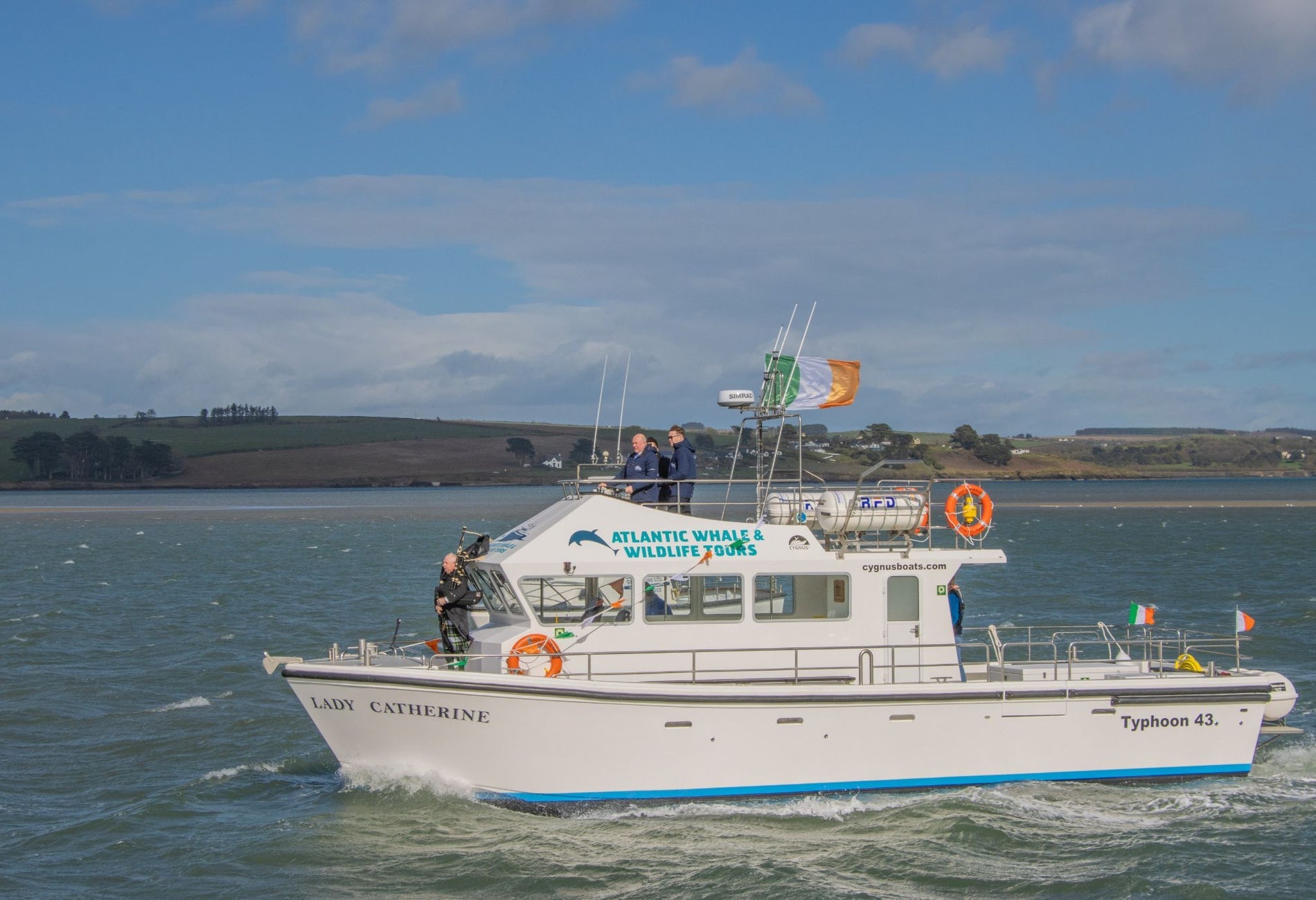 Three people on a white boat in Kinsale Harbour with the Irish flag flying