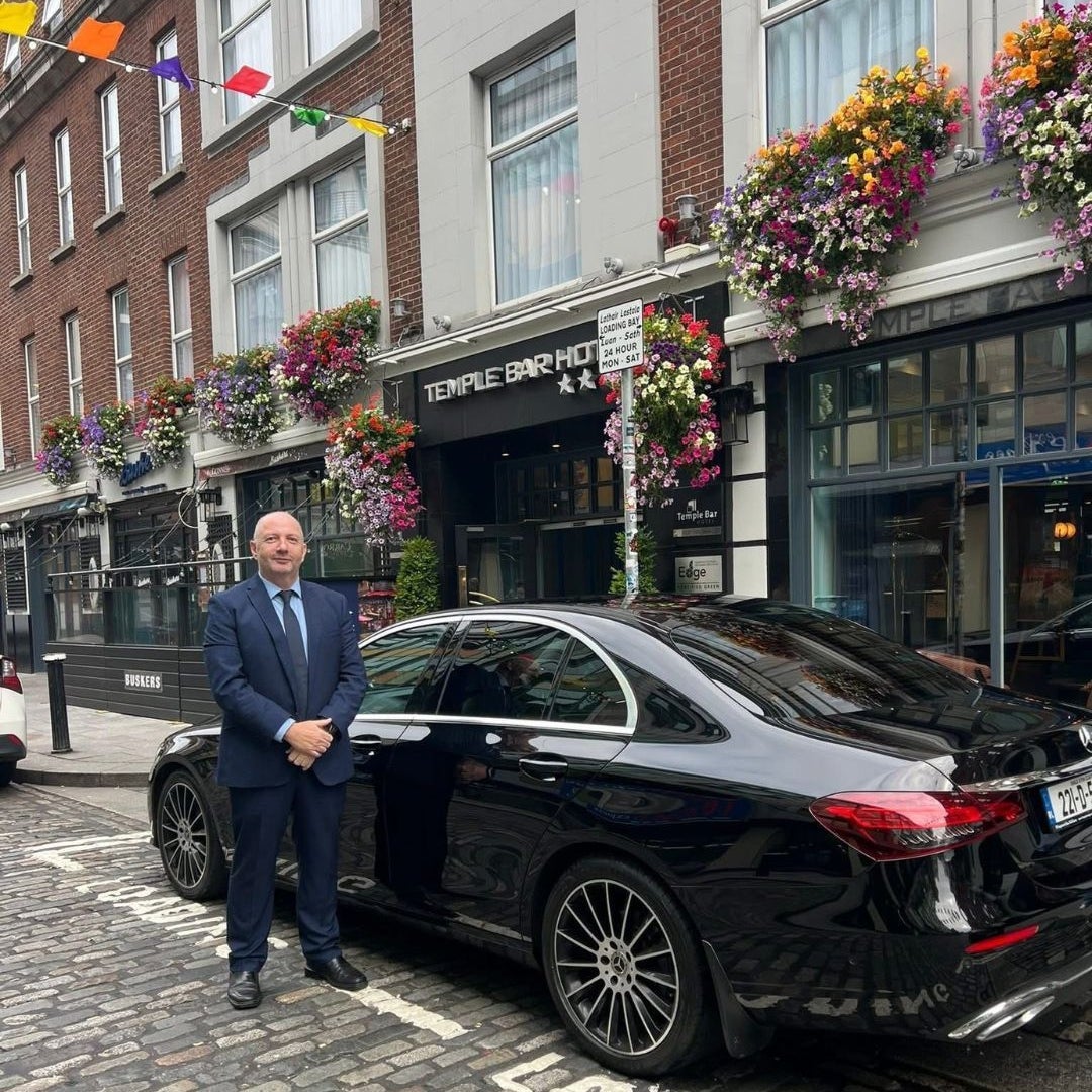 Person in a suit standing next to a black car parked on a street outside a hotel with flowers