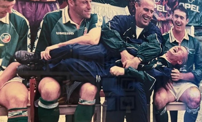 A group of laughing men in football strips seated with another man laid horizontally across their laps also laughing.