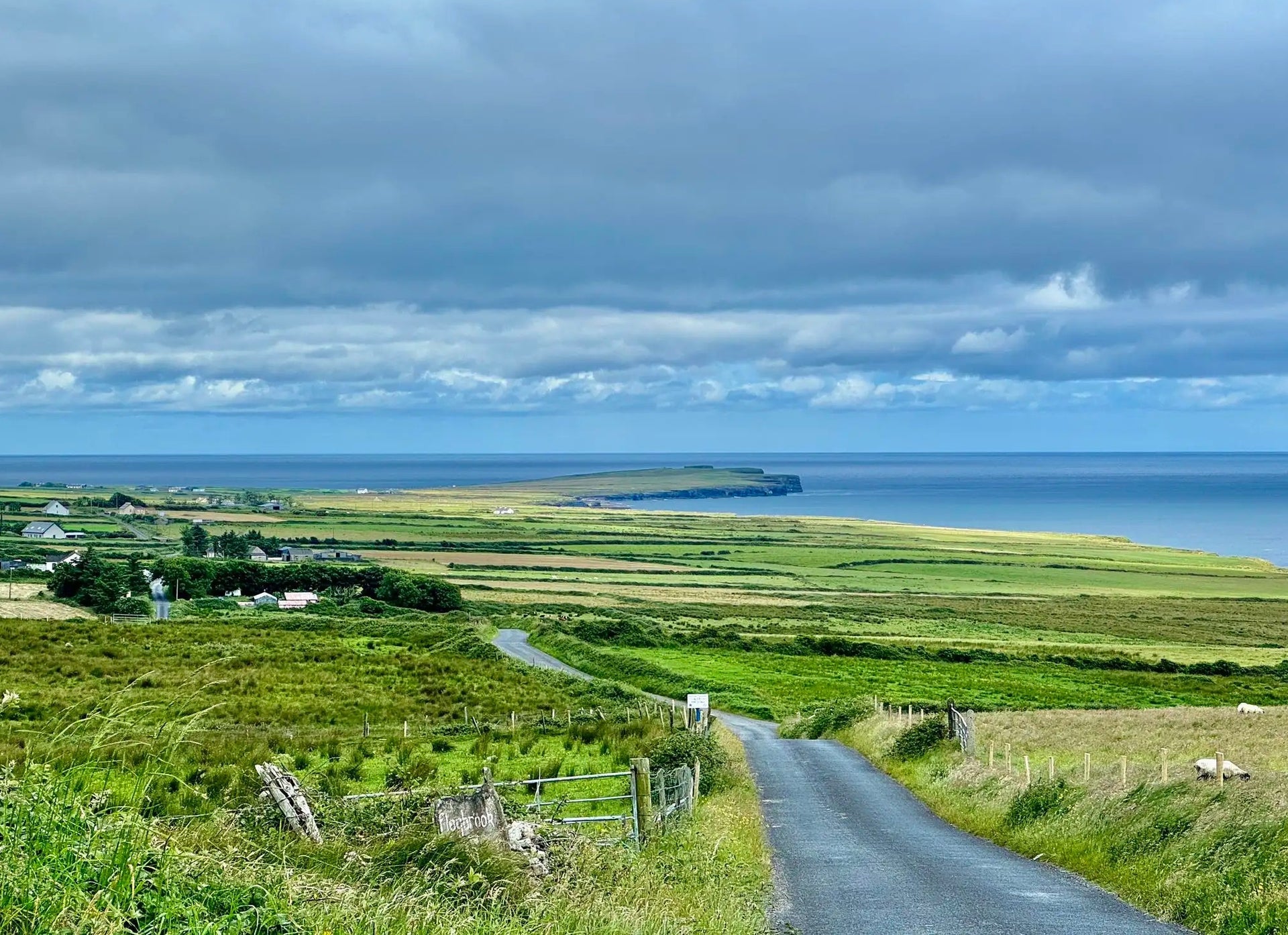 A country road meanders through green countryside with a cloudy sky and the sea in the distance