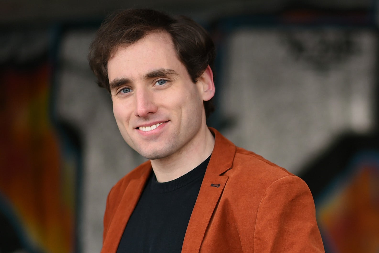 A man wearing a dark top and brown jacket is smiling at the camera.