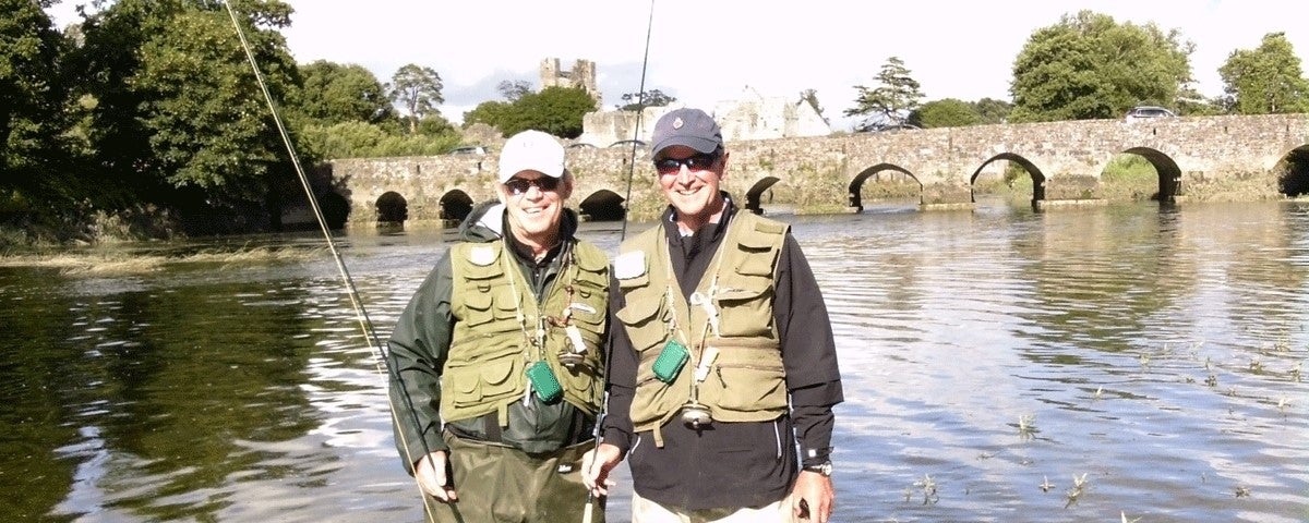 Two fishermen standing in the water holding fishing rods with a bridge in the background