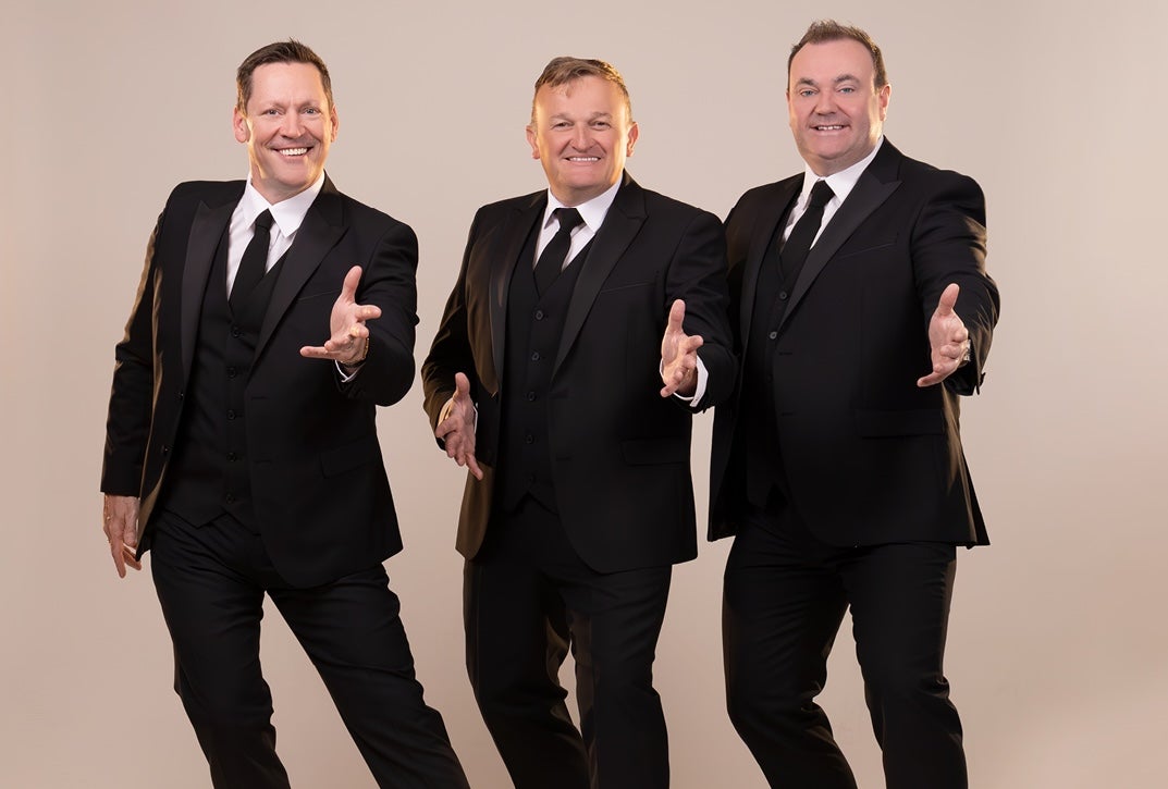 3 smiling men in black suits and ties each with their left hand outstretched, against plain beige background.