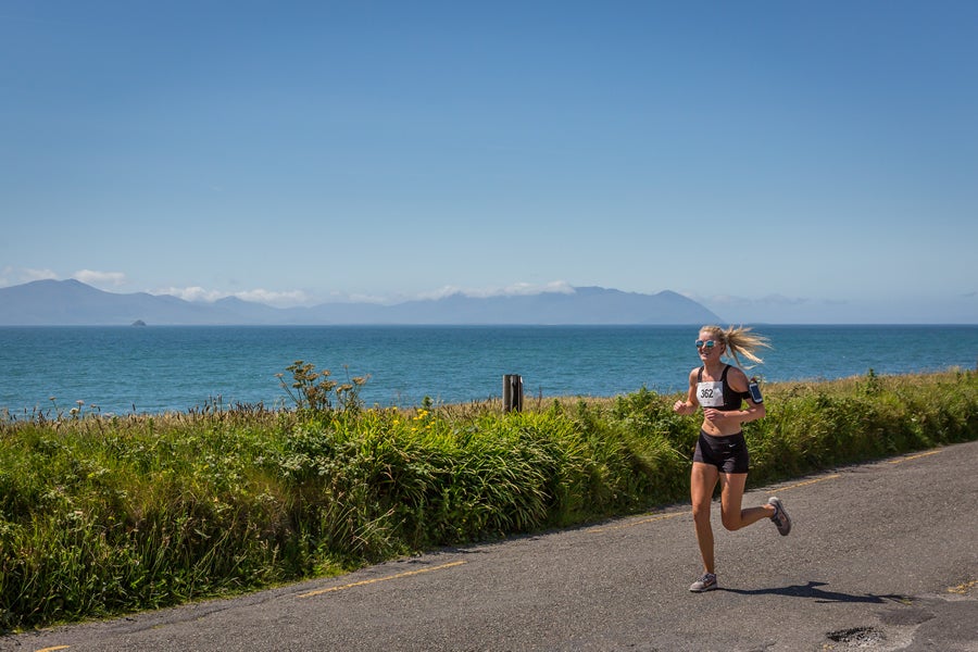 A sole figure running along a road with water and distant hills in the background and clear blue sky.