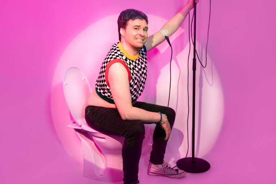 A grinning person seated, holding onto a mic on a stand with a bright pink background.
