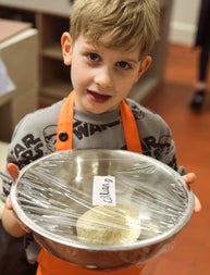 Child in an orange apron holding a silver bowl with a ball of dough in it