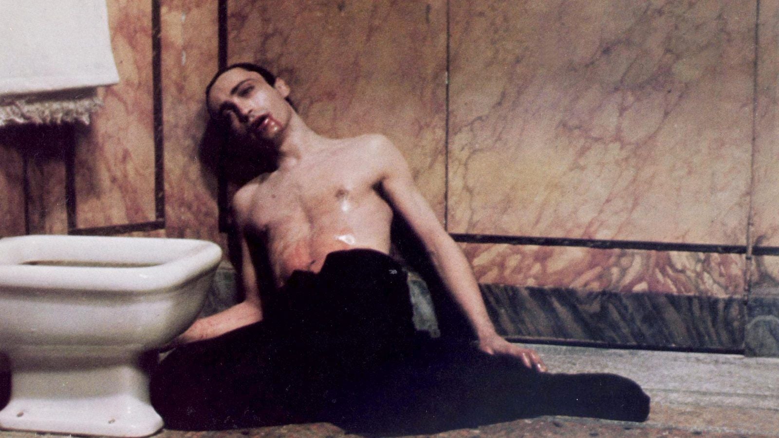 A shirtless man with waxy skin, with blood dripping from his mouth, is lying against a wall next to a toilet.