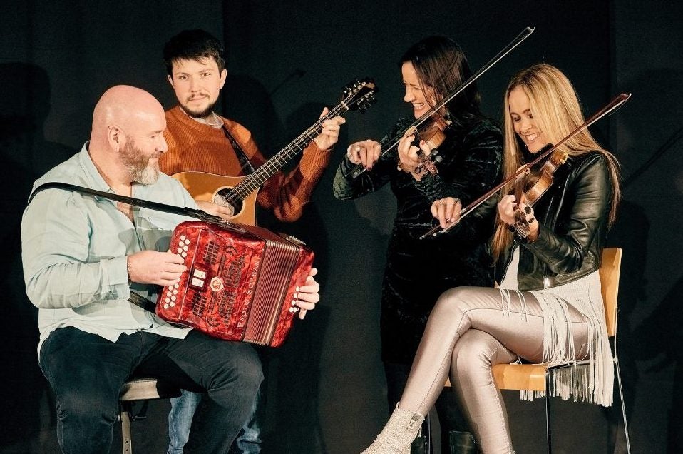 4 smiling musicians with their instruments, 2 seated with 2 standing behind, against plain, dark background.