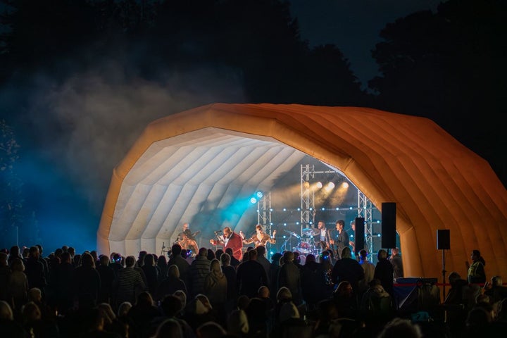 A blow up outdoor stage in orange with blue and white lights on performers at night with audience in darkness.