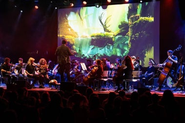 Orchestra performing on stage with strings in the foreground and the conductor, large screen behind showing gameplay footage from the game. Audience silhouettes in foreground.