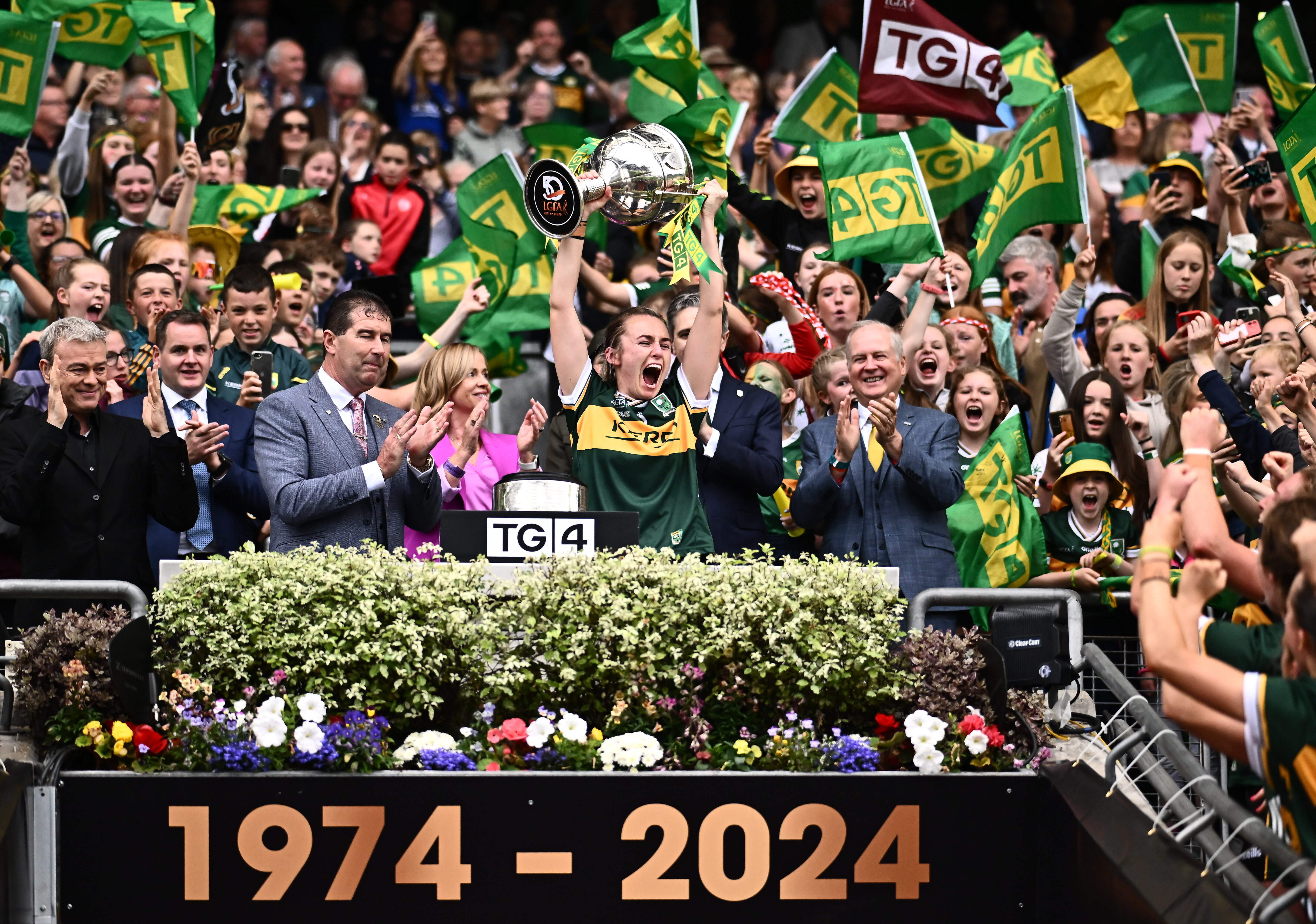 Celebrations in Croke Park, a sports person in jersey lifting large silver trophy over their head with clapping men either side in suits, against large crowd waving flags behind.