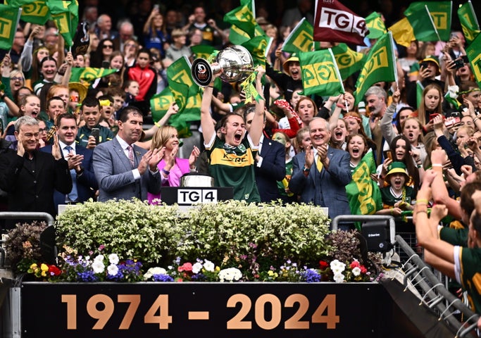 Celebrations in Croke Park, a sports person in jersey lifting large silver trophy over their head with clapping men either side in suits, against large crowd waving flags behind.