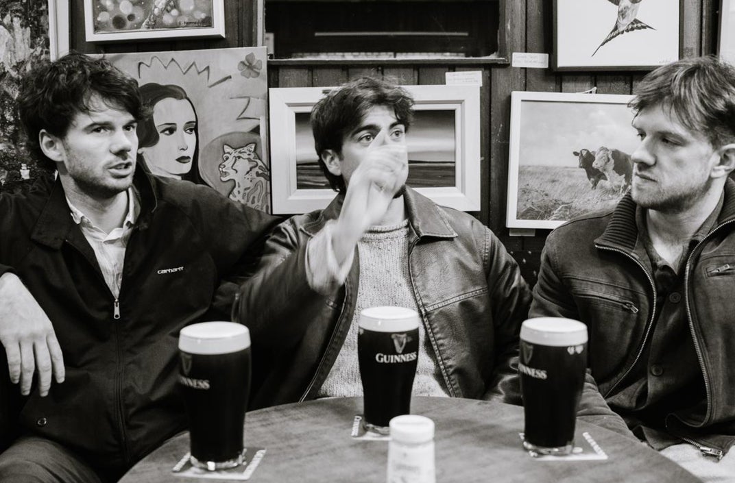 Black and white photo of 3 men sitting around a table with full pints of stout.