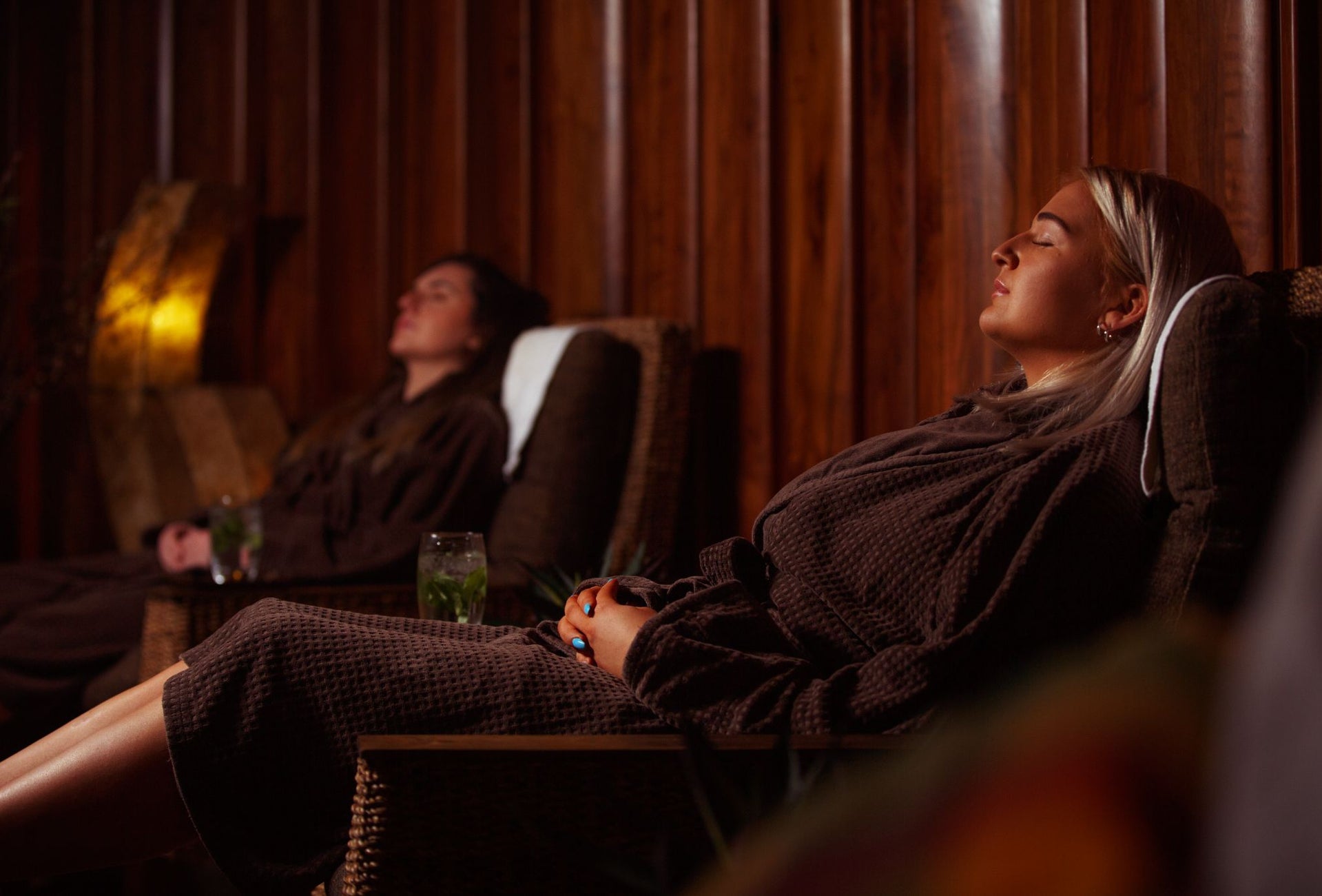 Two ladies in dressing gowns relax with a healthy drink on lounge seating in a spa