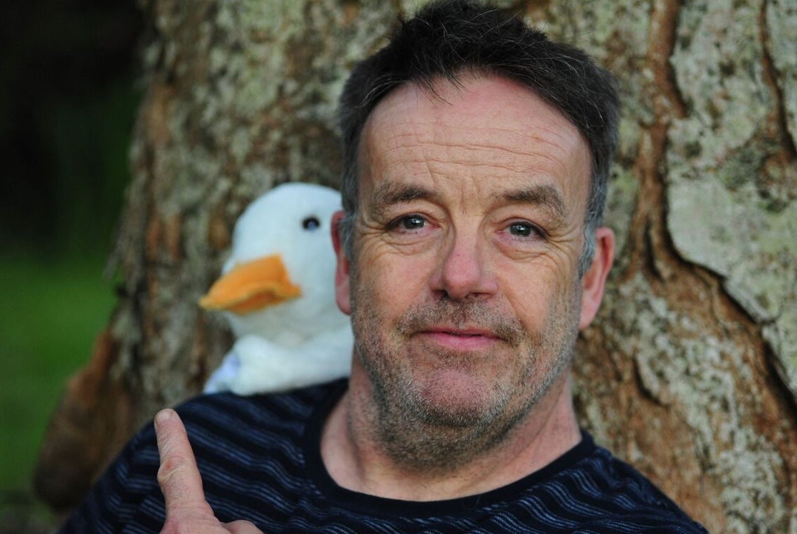 Púca – John Colleary: Mighty Quack, a close up of a man pointing over his shoulder at a white toy duck behind him