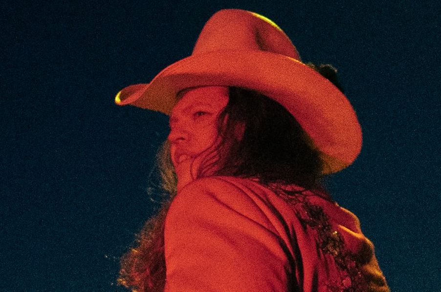 Slightly grainy red lit photo of person in a cowboy style hat and long hair, viewed from the rear, looking over their left shoulder, against a black background.
