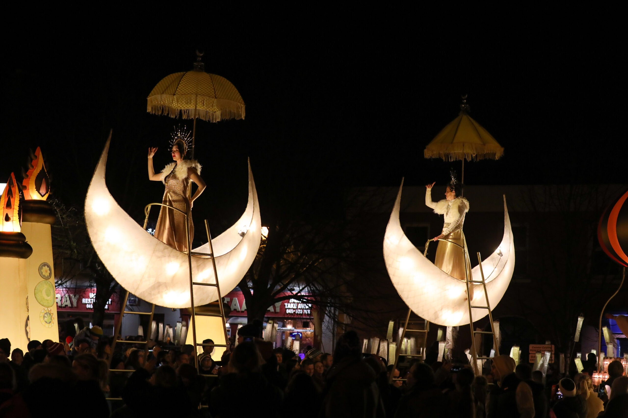 at night outdoors crowds are looking up at 2 illuminated half moon shapes each containing a person posing in a gold costume