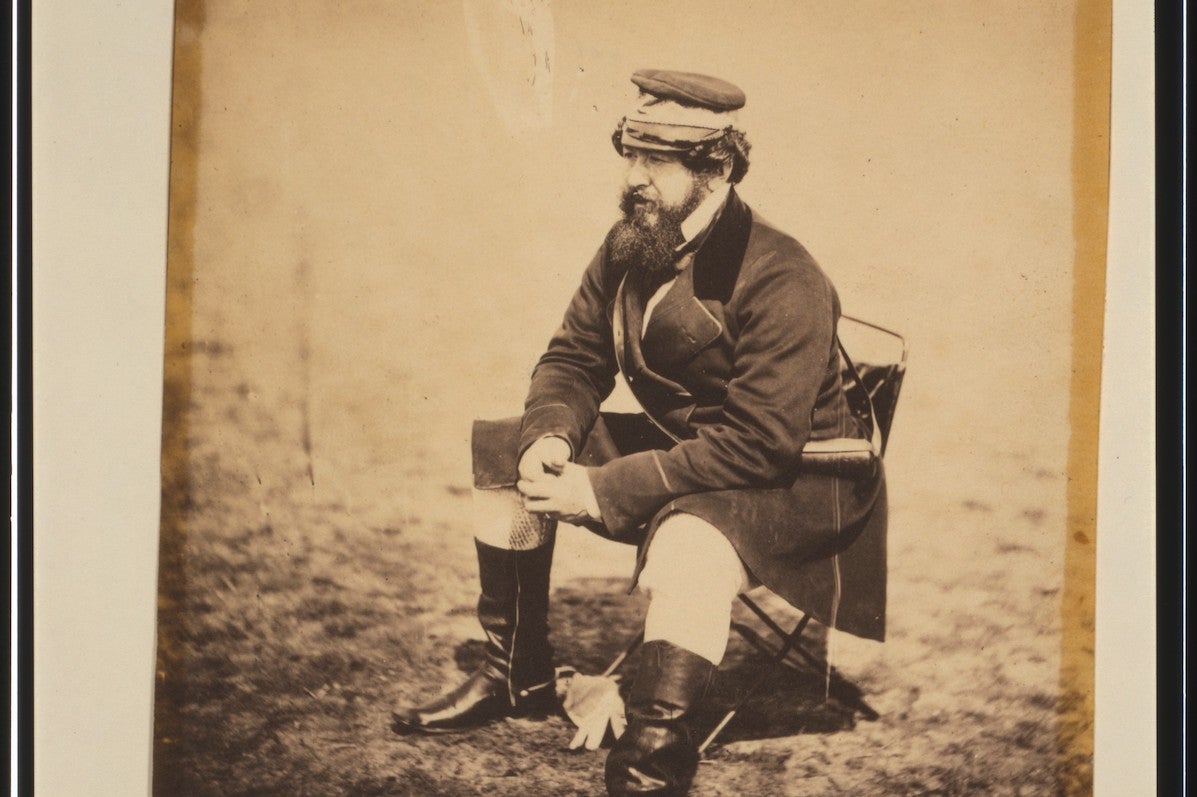 An old, brown style photo of a man in uniform with black boots seated on a camping chair.