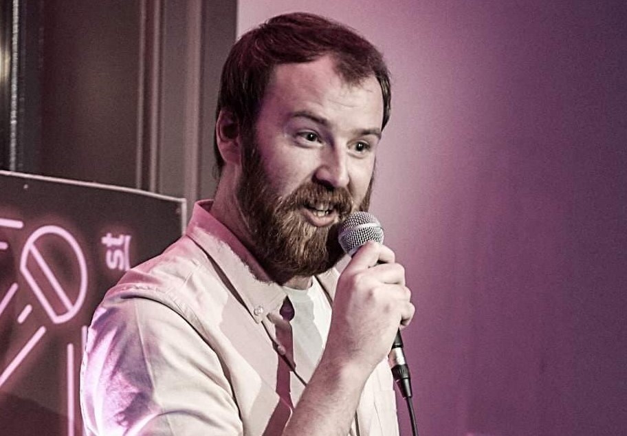 A man with a beard is talking into a mic he's holding, against purple background.