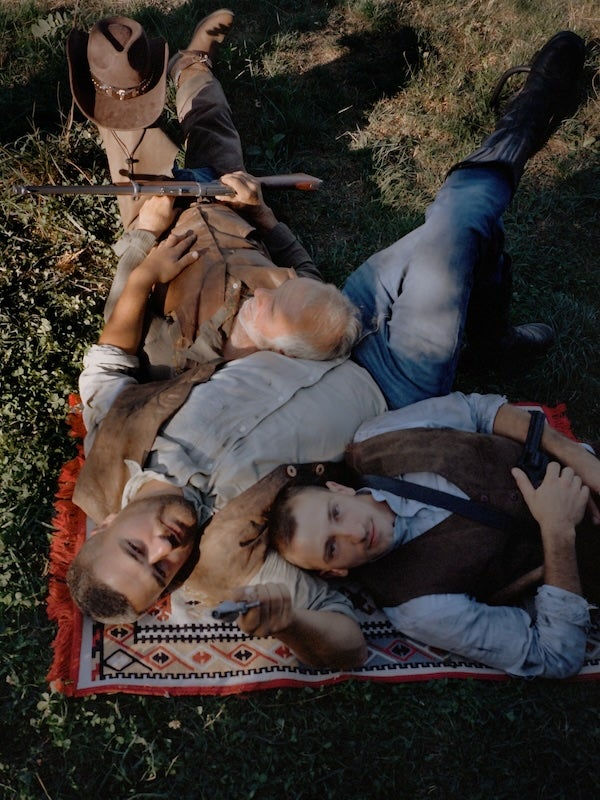 A photograph taken from standing height of three men dressed in Western style clothing lying on a woven rug on a grassy terrain. The men are of various ages, each resting his head upon the other.